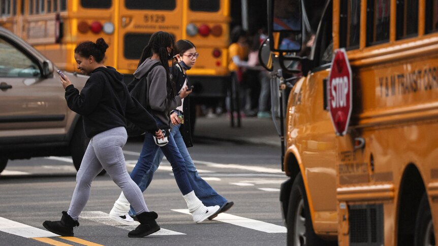 Children walk across a pedestrian crossing past multiple yellow school buses. 