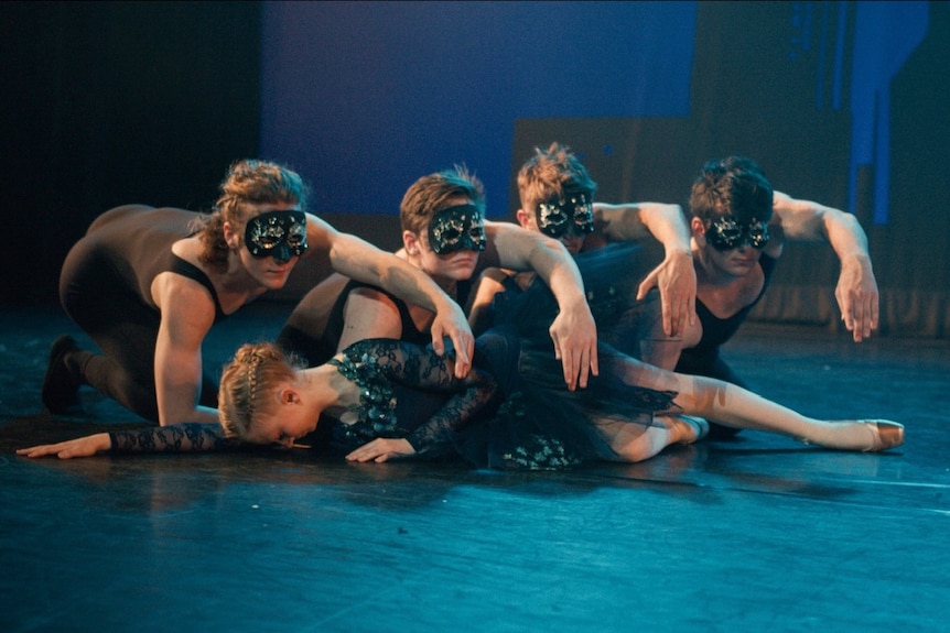 ballerina laying on the stage with male ballerinas leaning over her in black masks