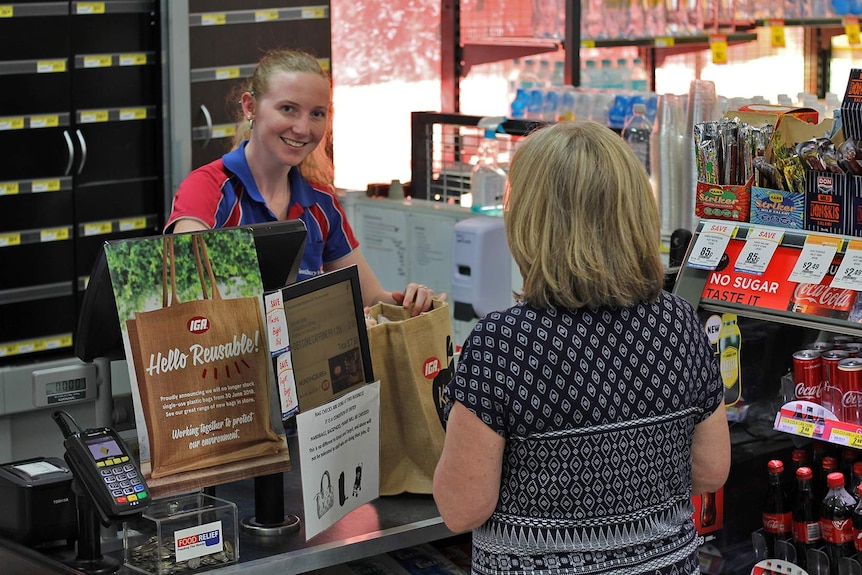 A young women serves behind the counter of a supermarket, she puts the customer's groceries into a reusable hessian bag.