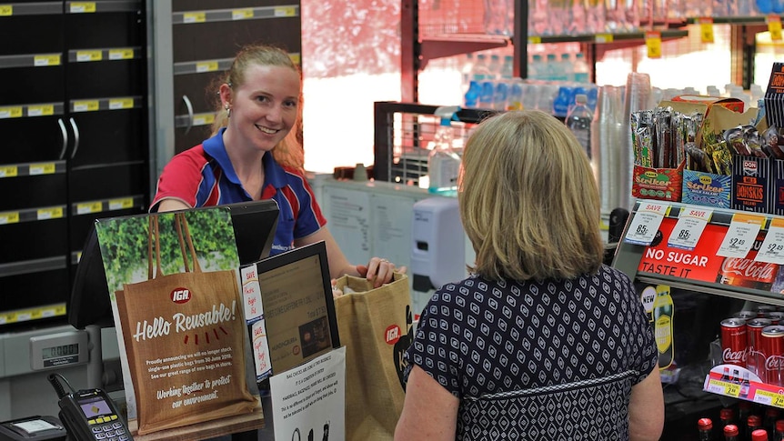A young women serves behind the counter of a supermarket, she puts the customer's groceries into a reusable hessian bag.