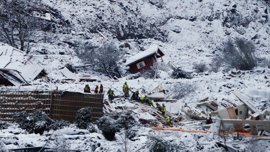 People in high-vis vests crawl over the rubble of a destroyed house in an icy and rocky landscape.