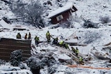 People in high-vis vests crawl over the rubble of a destroyed house in an icy and rocky landscape.
