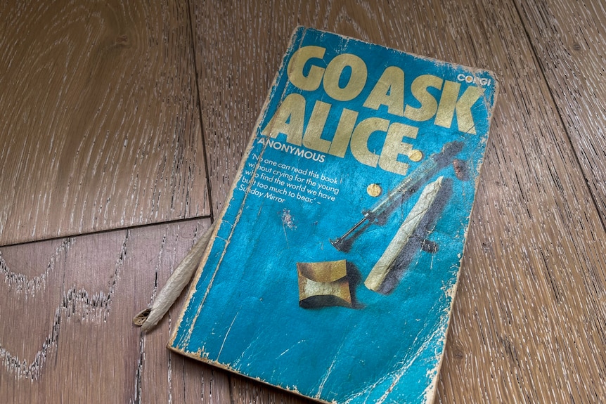 A tattered book sits on a floor. The cover is blue, with drug paraphernalia and GO ASK ALICE - ANONYMOUS written on the front