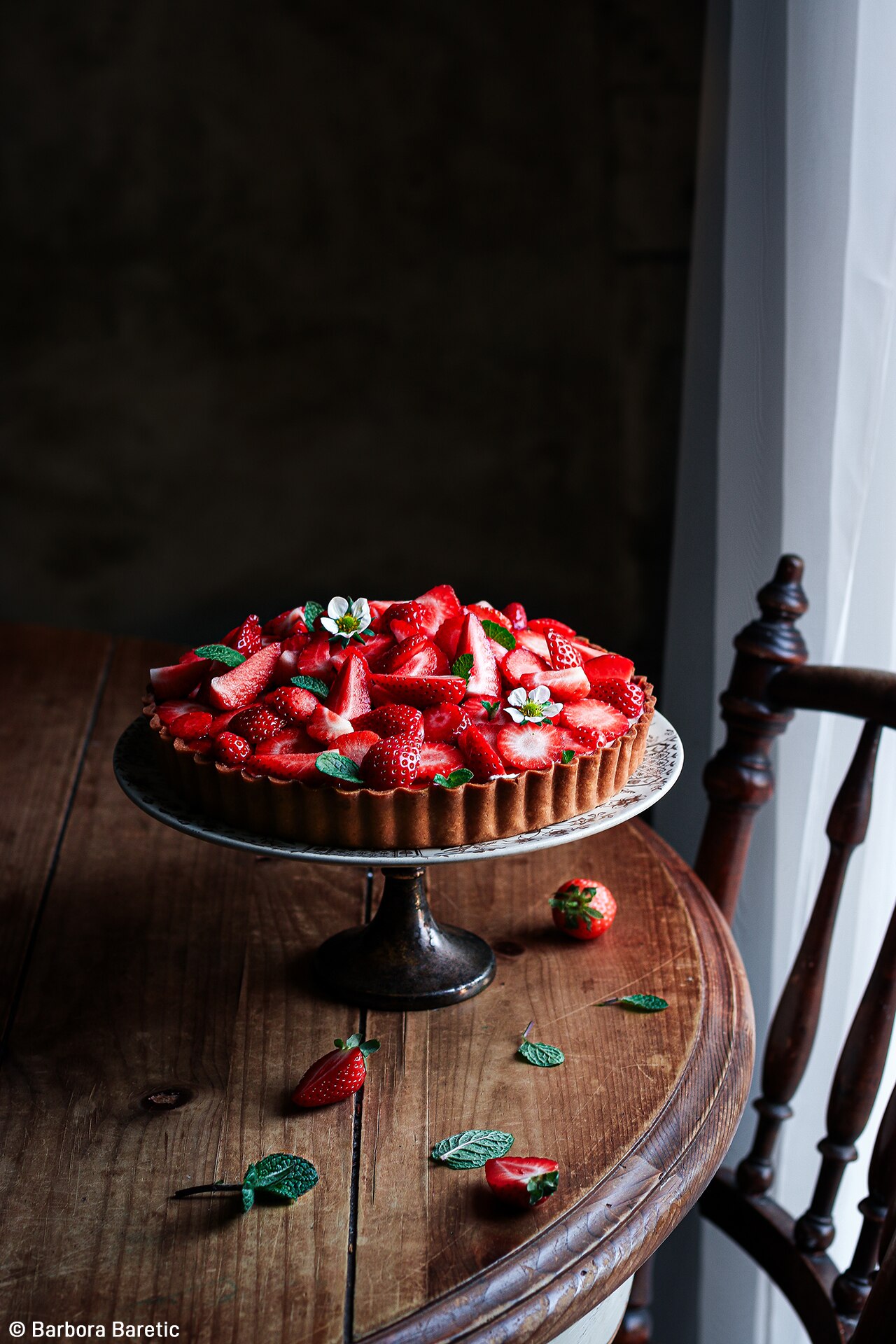 A tart covered in strawberries sitting on a cake stand, on a table. 