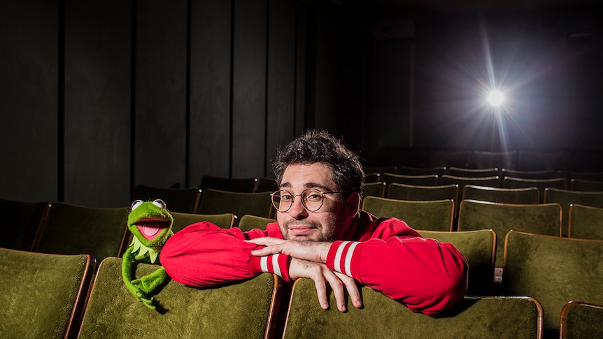 Artist David Capra and a puppet Kermit the Frog leaning forward on cinema seats in a darkened cinema.