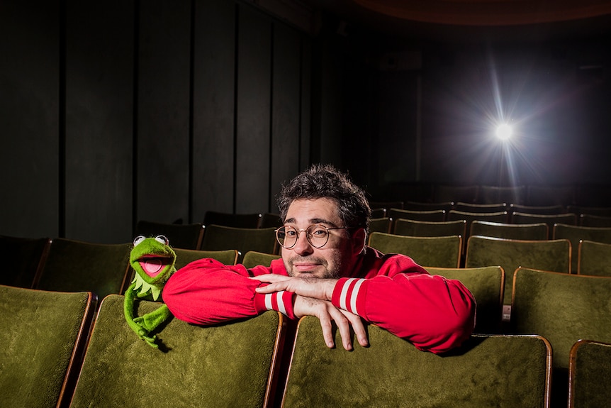 Artist David Capra and a puppet Kermit the Frog leaning forward on cinema seats in a darkened cinema.