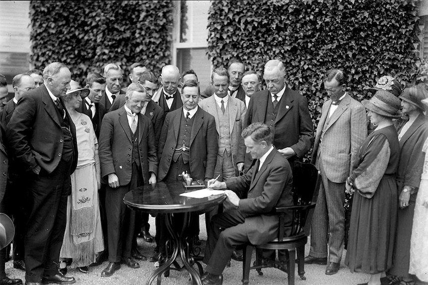 A man is seated signing a document in front of a crowd of finely dressed men and women in a garden
