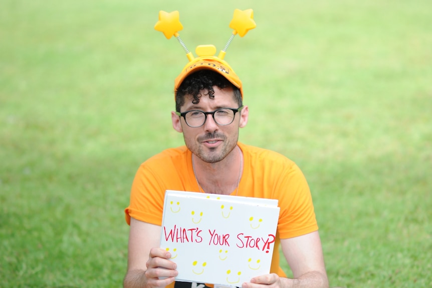 Caleb Rixon is dressed in a yellow t-shirt and hat as he sits holding a card reading 'what's your story' in a park.