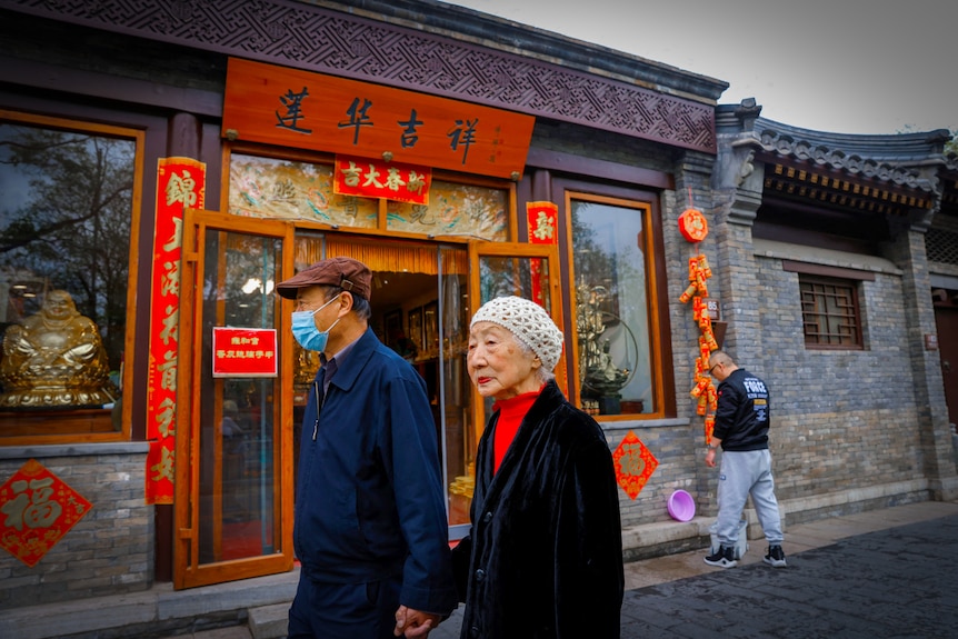 An older man in a face mask holds hands with an older woman as they walk past a temple in Beijing