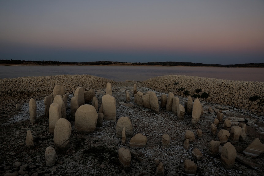 Dozens of rounded stones rise from a rocky lake bed against a sunset horizon.