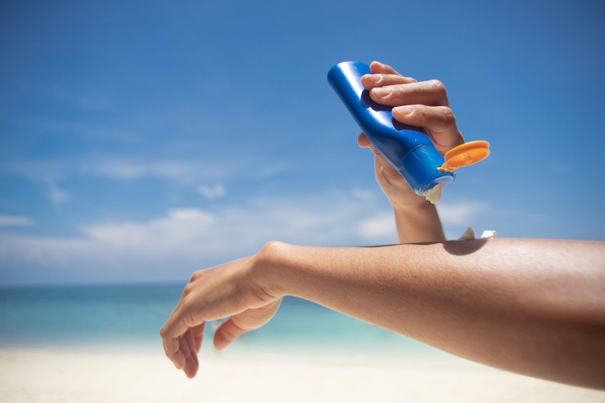A person at the beach applying sunblock to their arm. 