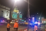 Police line blocking off a road with a bus and yellow tarmac up. Tradies looking on. 