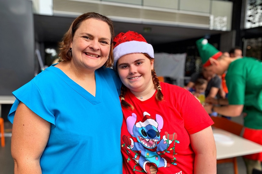A woman in a blue shirt hugging a smaller woman in a red shirt and santa hat smiling