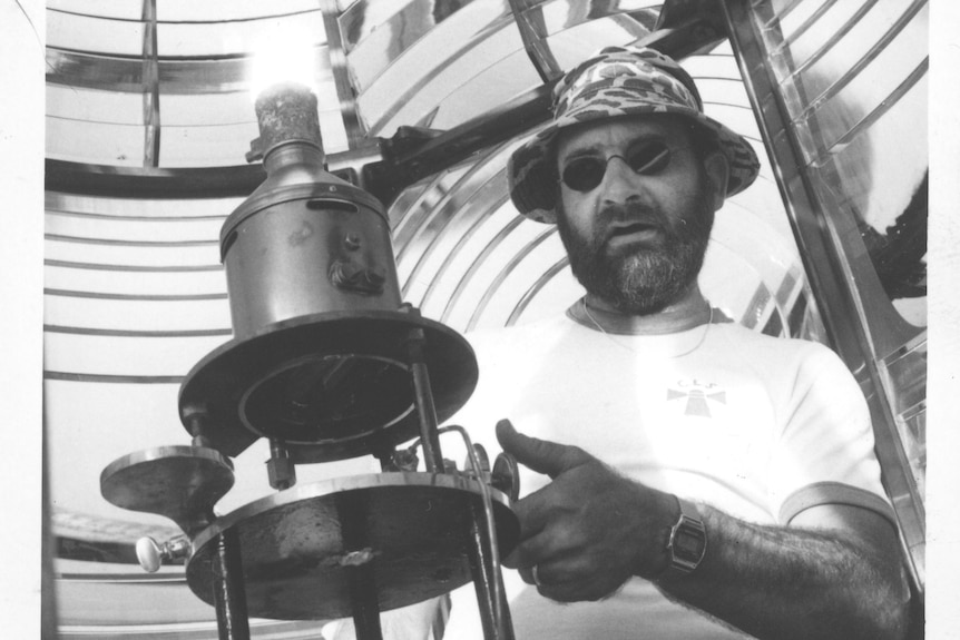 Black and white photo of man with sunglasses on standing in front of a kerosene light mantle inside a lighthouse.