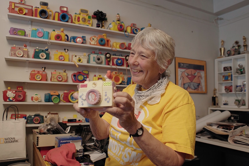 A smiling woman wears yellow t-shirt smiles and holds up toy camera in front of wall shelves full of toy cameras in apartment.