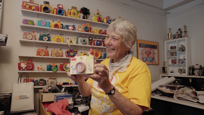 A smiling woman wears yellow t-shirt smiles and holds up toy camera in front of wall shelves full of toy cameras in apartment.