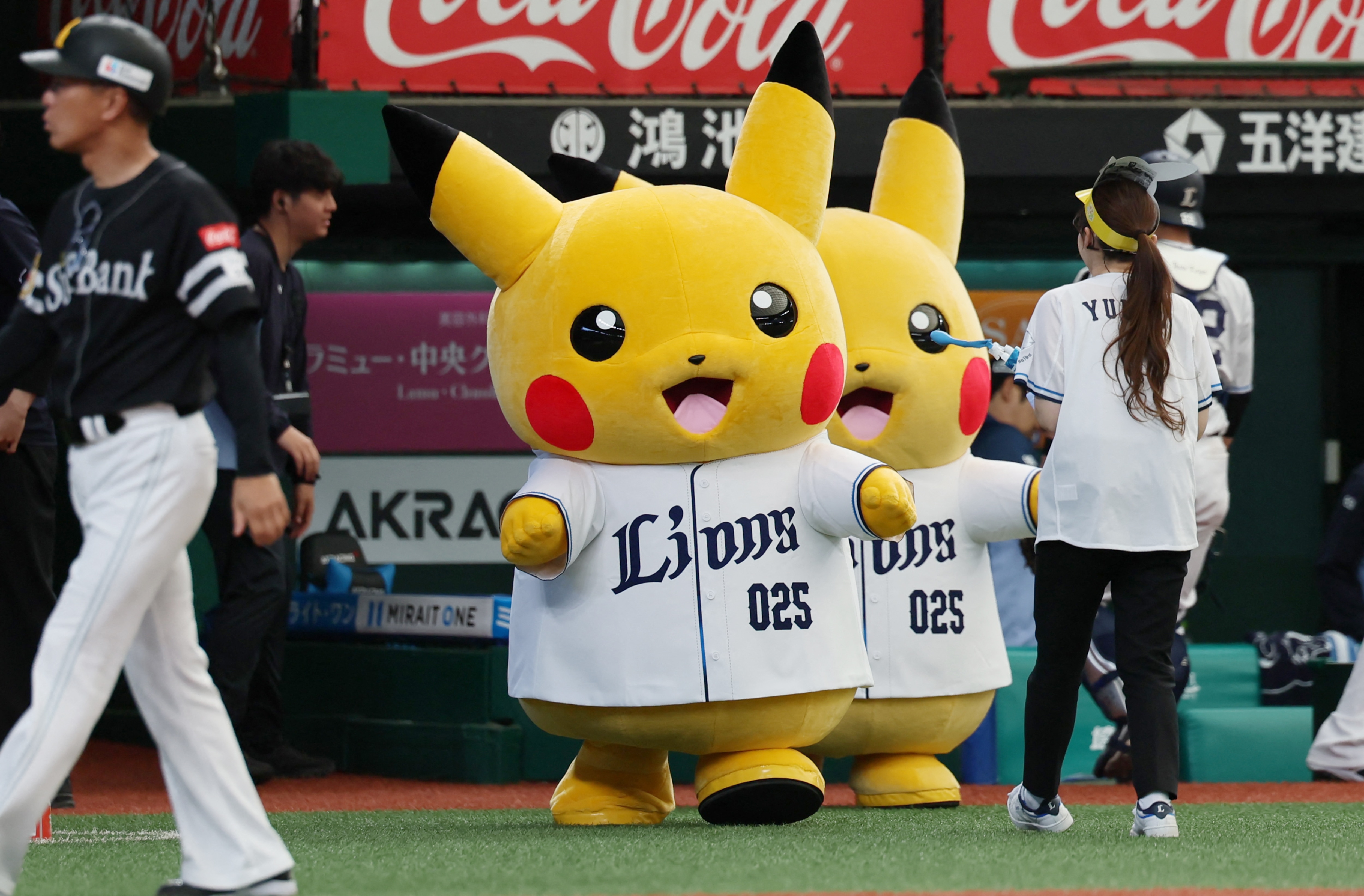 Two Pikachu mascots on a baseball field as a player walks past.