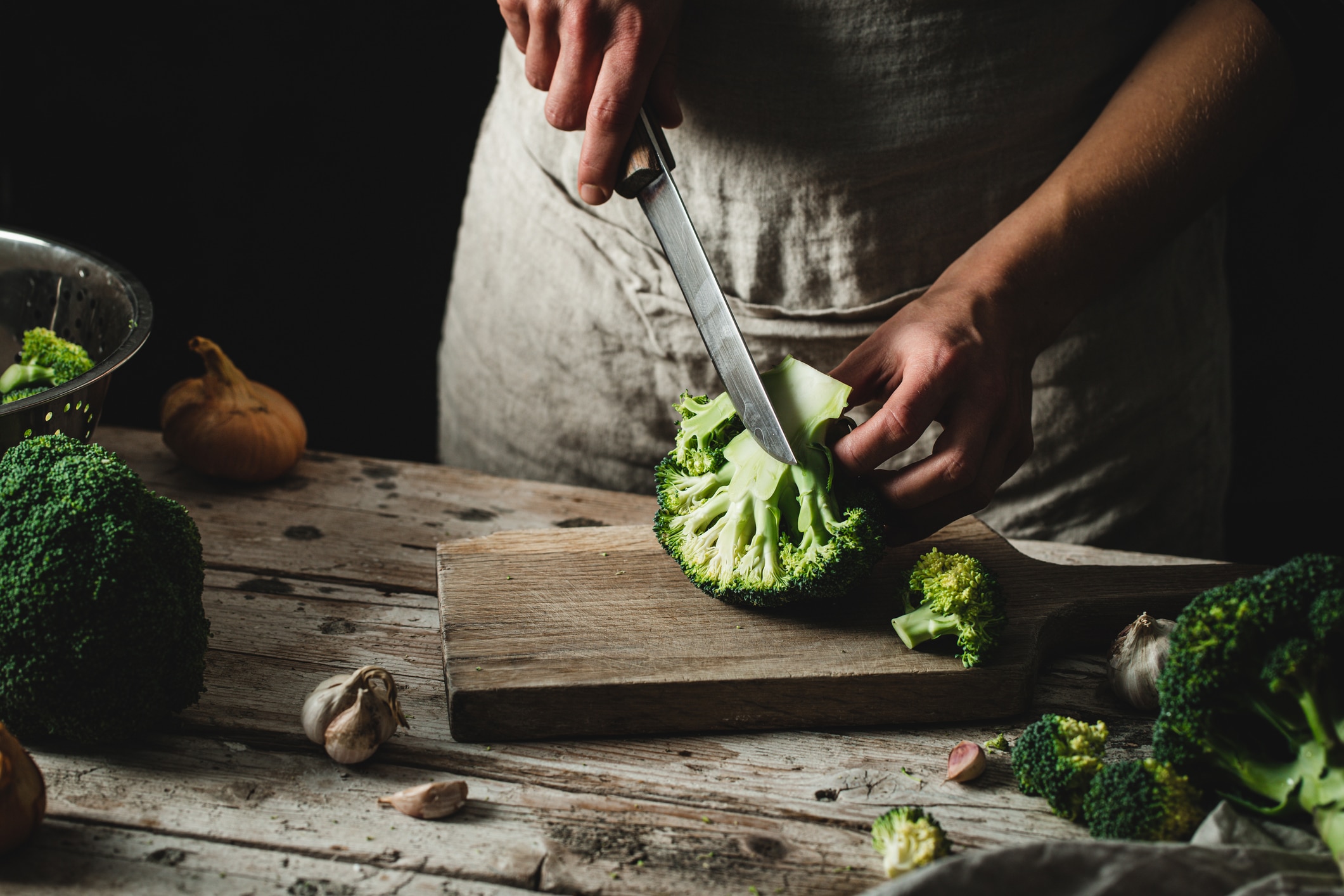 In a darkly-lit scene, you view a woman finely slicing a broccoli on a wooden board with a sharp knife.