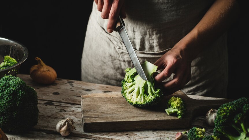 In a darkly-lit scene, you view a woman finely slicing a broccoli on a wooden board with a sharp knife.
