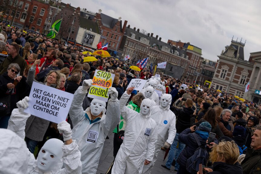 Protesters in white masks and jumpsuits walk in a line through a crowd in a city square.