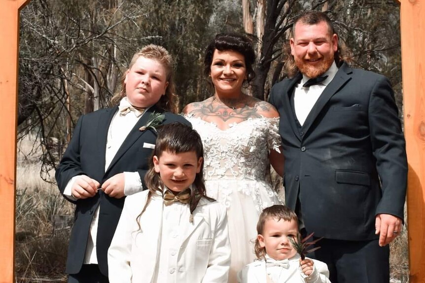 A family portrait with a woman wearing a white wedding dress and the boys suits, all have mullet hairstyles.