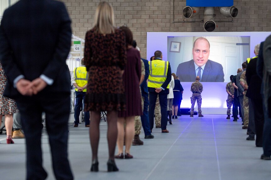 Prince William on a screen with people standing and watching while socially distancing.
