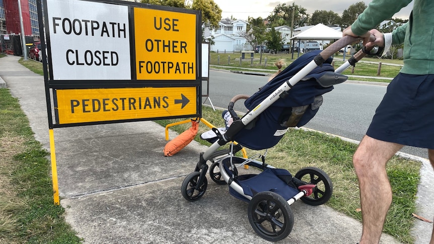 Man standing with a pram on a closed footpath with a yellow detour sign.