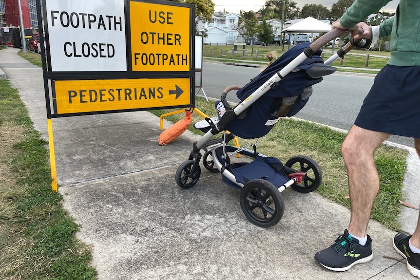 Man standing with a pram on a closed footpath with a yellow detour sign.