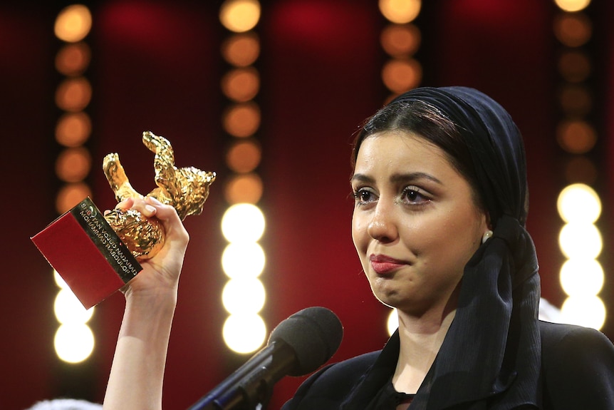 A 20-something-year-old Iranian woman wearing black head scarf stands at microphone looking emotional, holding golden statuette.