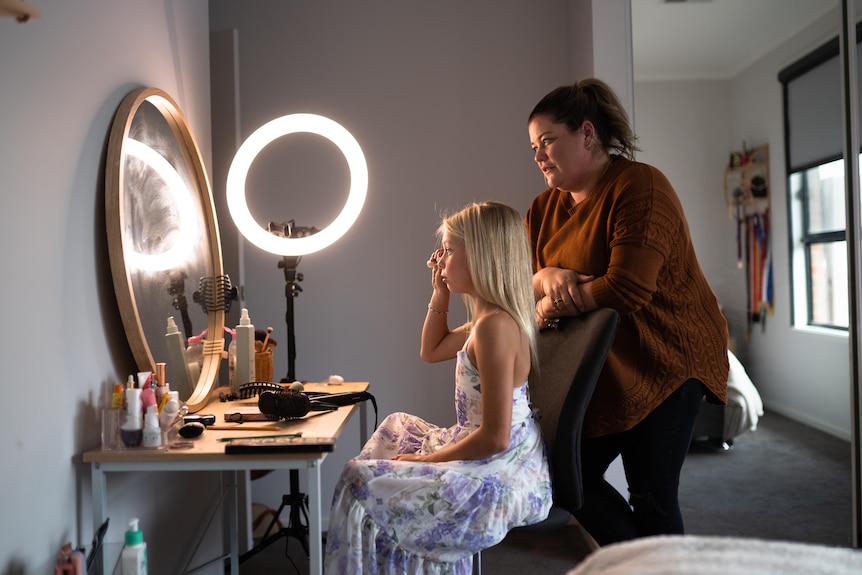 A young girl with blonde hair applies make-up in front of a mirror lit with a ring light, while a woman watches on.