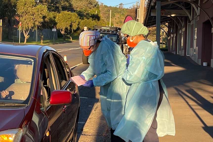 Two people wearing full protective equipment approach a car to perform a covid-19 swab