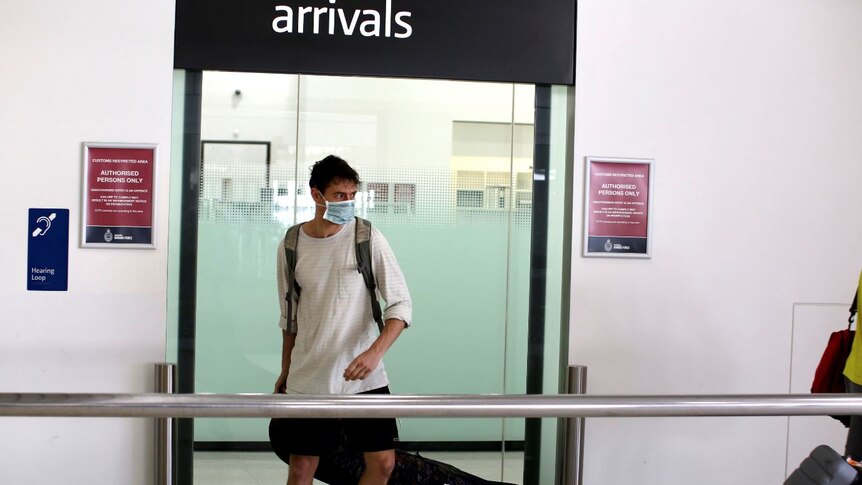 A man wearing a mask and pulling a suitcase walks under an arrivals sign.