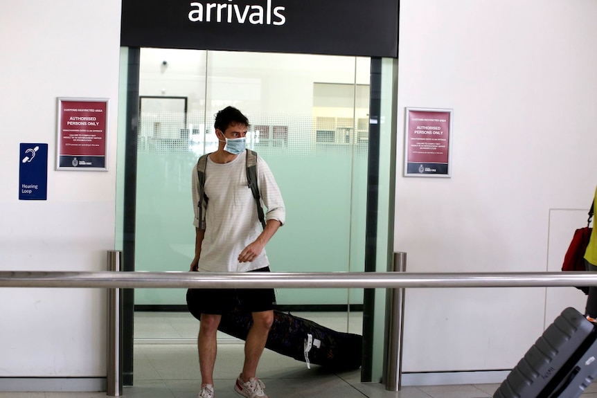A man wearing a mask and pulling a suitcase walks under an arrivals sign.