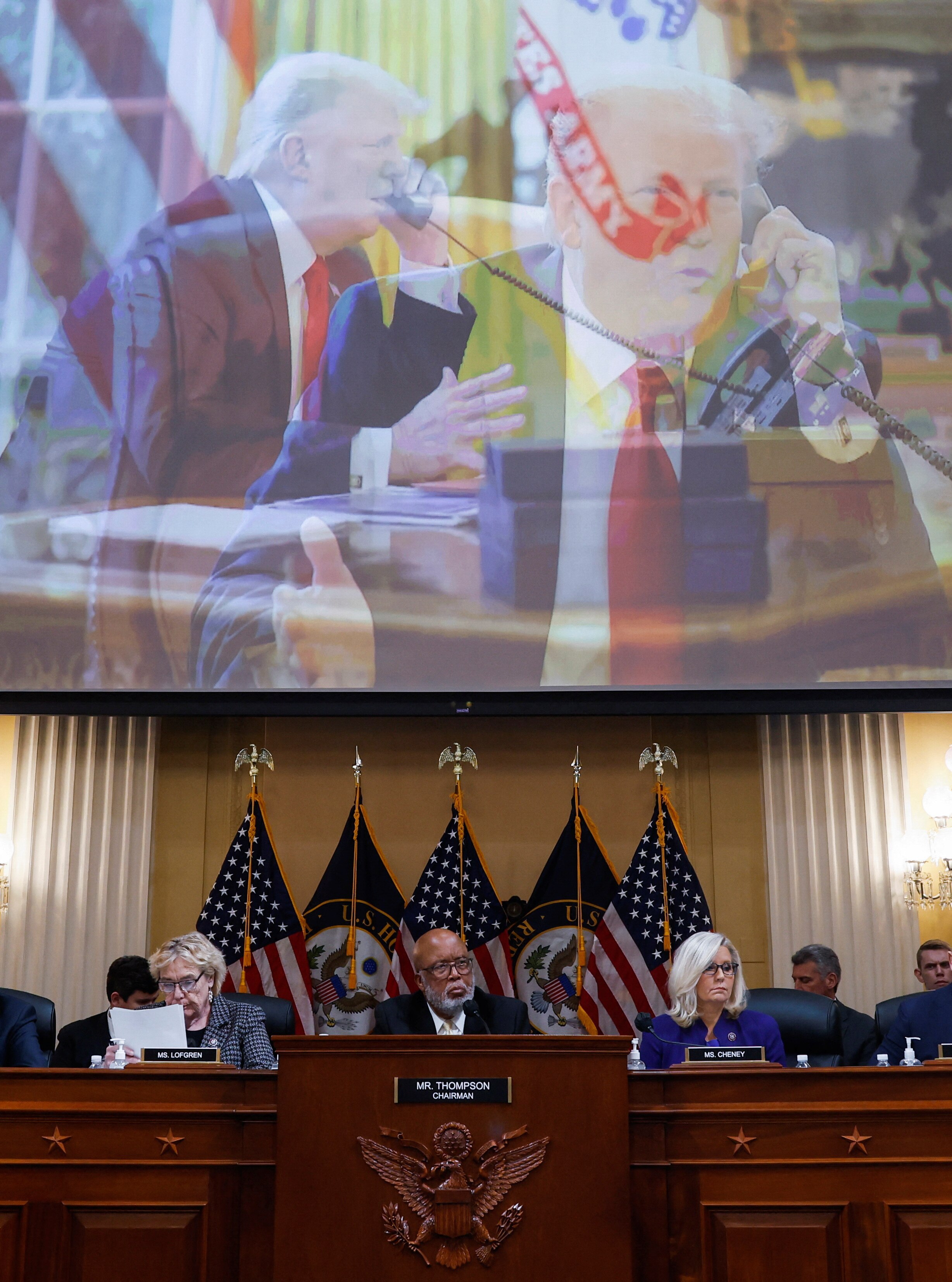 Zoe Lofgren, Bennie Thompson and Liz Cheney sit at a desk with a screen above them displaying Donald Trump