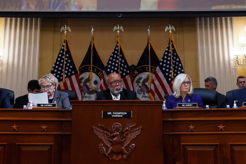 Zoe Lofgren, Bennie Thompson and Liz Cheney sit at a desk with a screen above them displaying Donald Trump