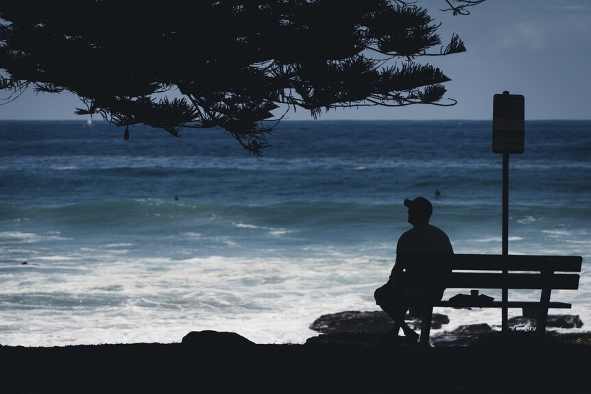 A man in silhouette sits at a bench and stares out at the ocean.