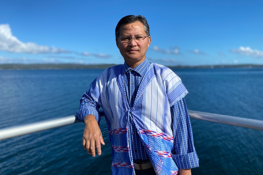 Karen man standing on jetty with rolling hills and water behind him wearing bright blue shirt