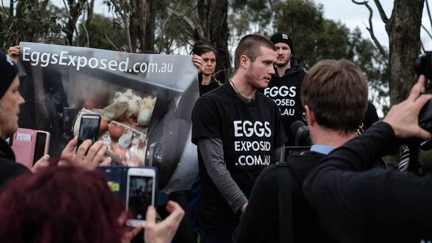 Chris Delforce stands in front of a crowd of journalists, with protesters holding banners behind him.