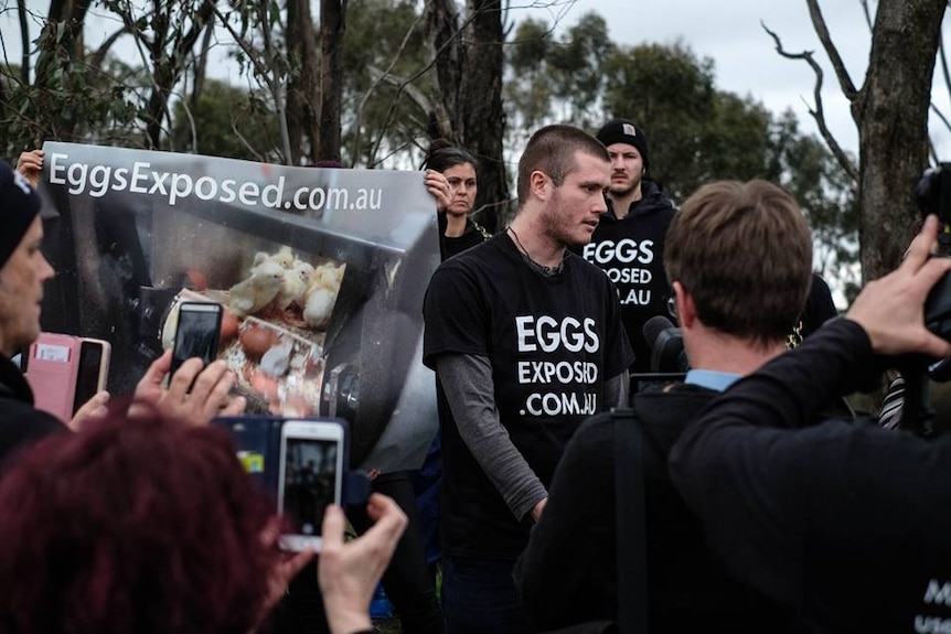 Chris Delforce stands in front of a crowd of journalists, with protesters holding banners behind him.