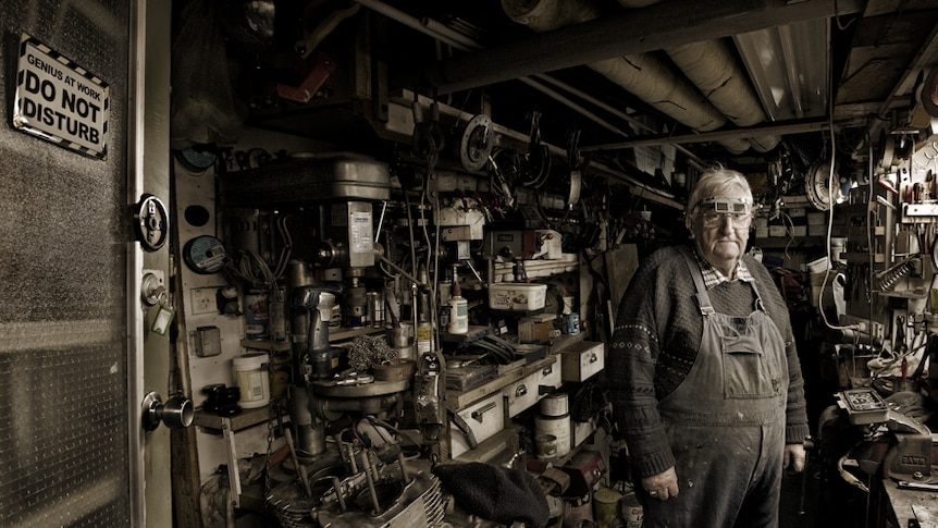 A man stand in a shed, workbenches crammed with tools on either side.