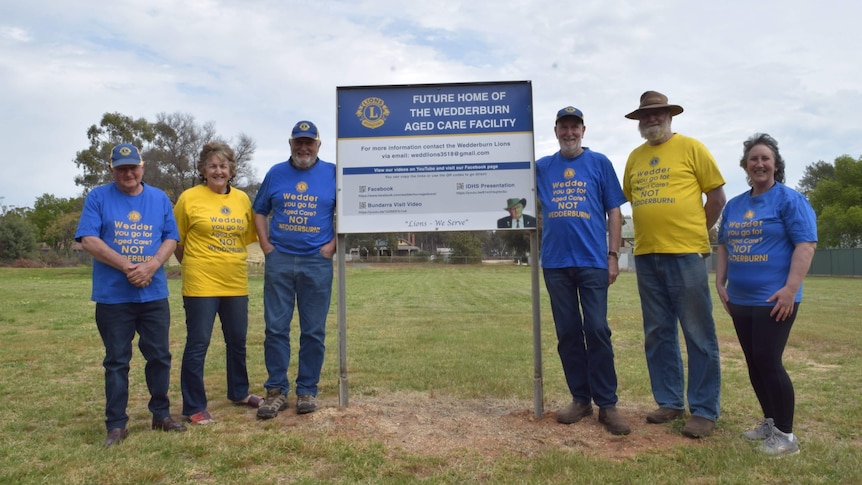 Six people in alternating yellow and blue t-shirts standing on a grassy field, beside a sign.