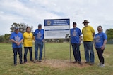 Six people in alternating yellow and blue t-shirts standing on a grassy field, beside a sign.