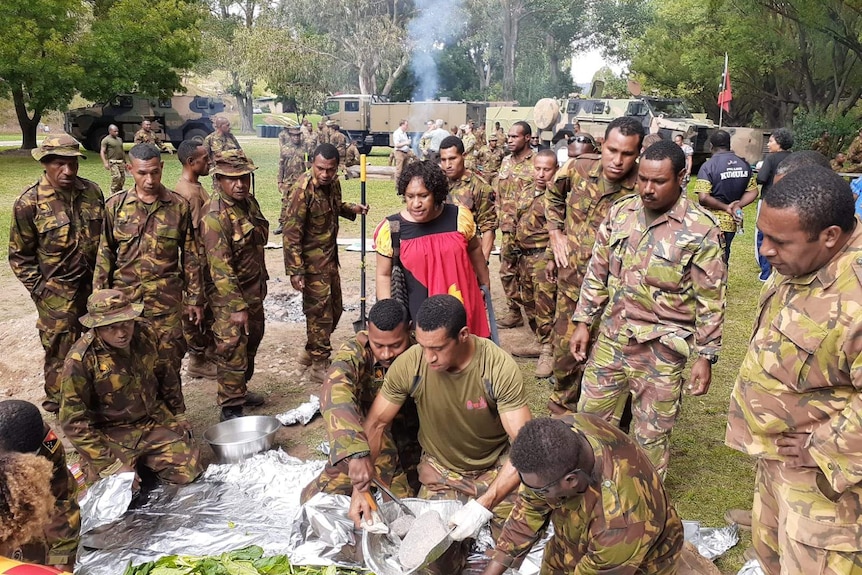 A woman stands surrounded by men in army fatigues preparing a large meal on the ground.