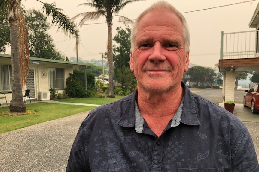 A man in a blue shirt with grey hair smiles at the camera, he stands in front of units in a smokey Mallacoota