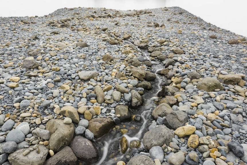A thin stream of water runs down a constructed riverbed of rocks, in a room with white walls
