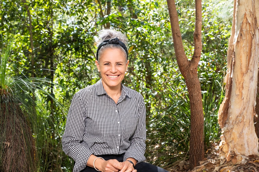 A woman in a button up shirt sits, smiling in between trees