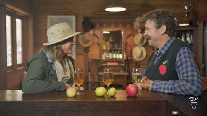 Man and woman sitting at a bar with apples and  glasses filled with amber-coloured liquid