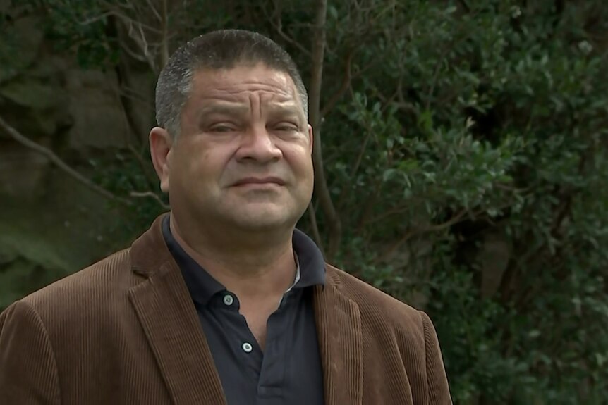 An Indigenous man wears a brown jacket and a serious expression
