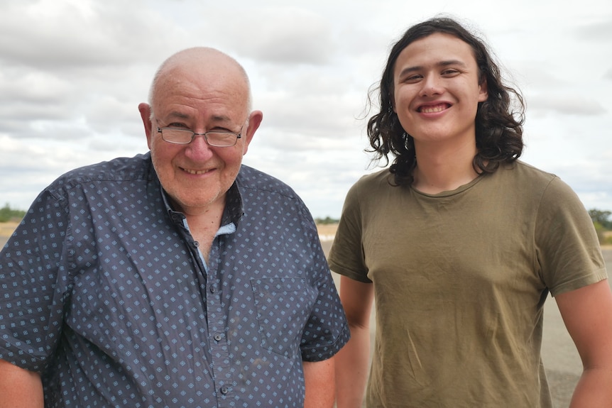 An older man stands next to his younger son with clouds behind them.