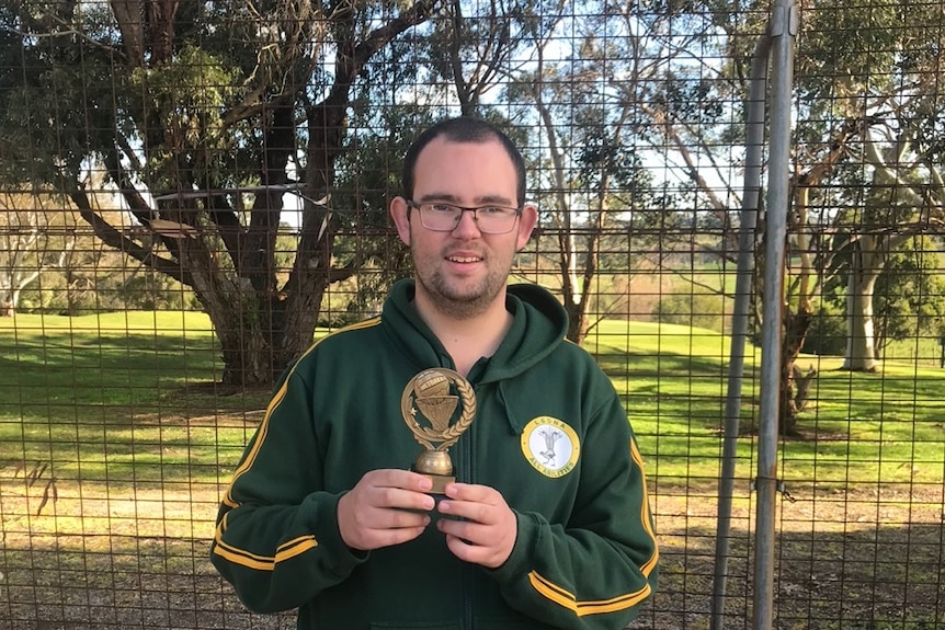 Damian wears a green hoodie holding a trophy in front of a wire fence with trees and grass in the background